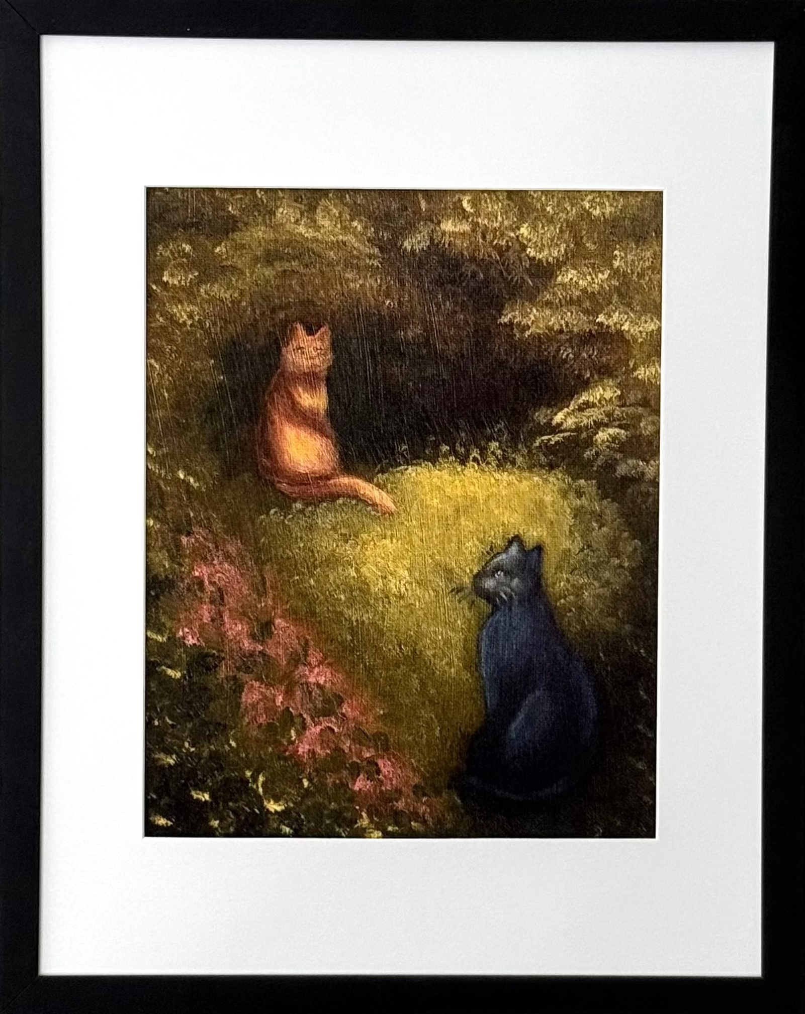 A framed art print featuring a painting of an orange cat and a black cat sitting in a grassy forest clearing, mounted on a light gray wall.