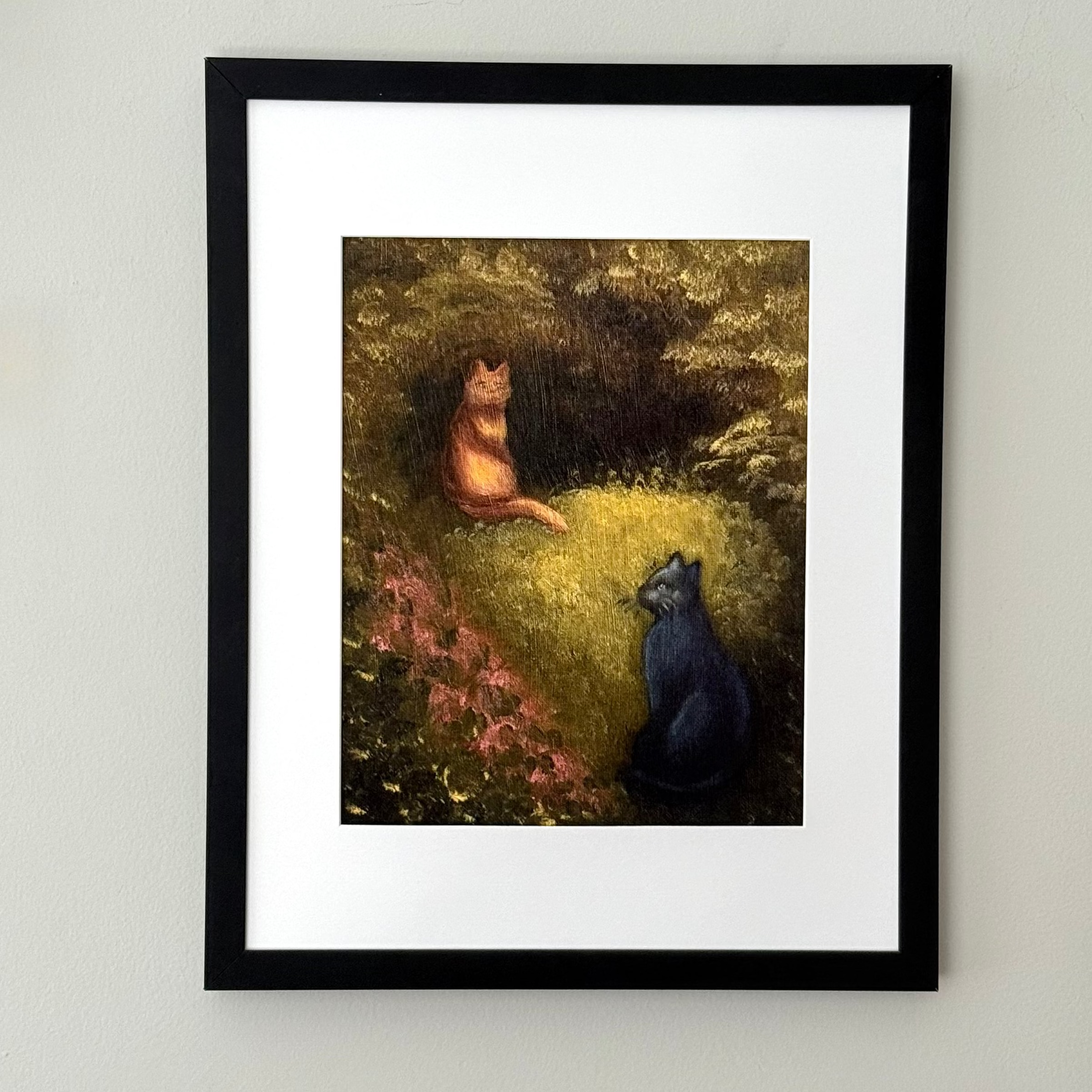 A framed art print featuring a painting of an orange cat and a black cat sitting in a grassy forest clearing, mounted on a light gray wall.