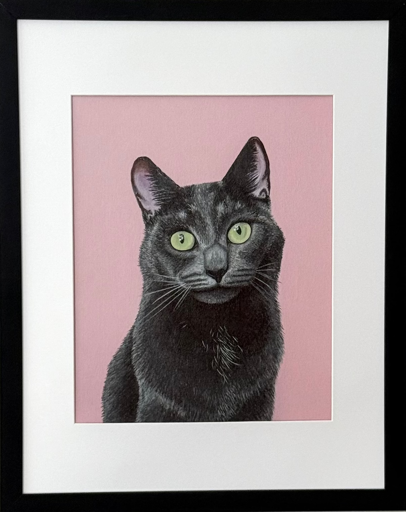 A framed art print featuring a black cat with green eyes on a pink background, mounted in a black frame with a white mat on a light gray wall.