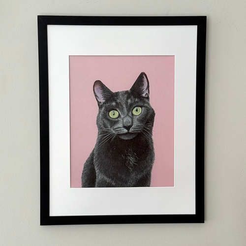 A framed art print featuring a black cat with green eyes on a pink background, mounted in a black frame with a white mat on a light gray wall.