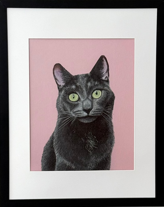 A framed art print featuring a black cat with green eyes on a pink background, mounted in a black frame with a white mat on a light gray wall.