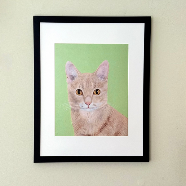 A framed art print featuring a portrait of an orange tabby cat on a light green background, housed in a black frame with a white mat, mounted on a beige wall.