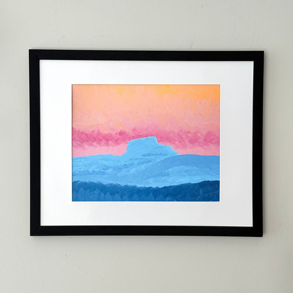 A framed landscape art print featuring a stylized sunset or sunrise with a pink and orange sky, blue mountains, and a dark blue foreground, displayed on a light gray wall.