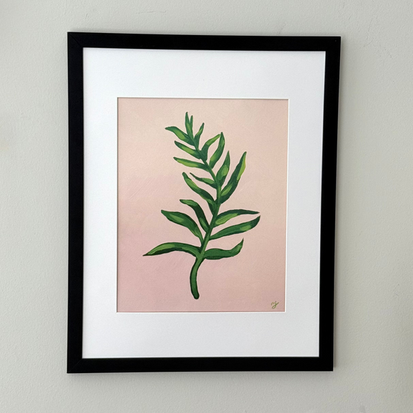 Framed botanical art print featuring a green leaf illustration on a soft pink background, matted with white and enclosed in a black frame, mounted on a light gray wall.
