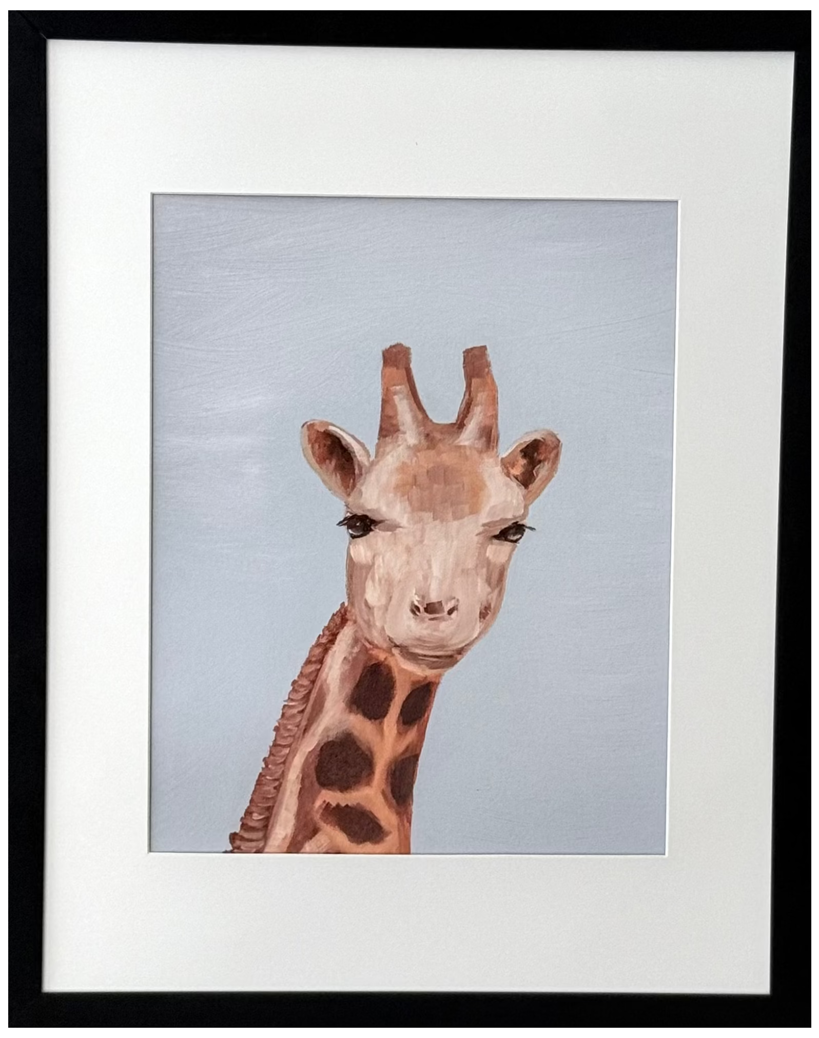 A framed art print featuring a painted giraffe head and neck against a pale blue-gray background, mounted on a light beige wall. The print has a white mat and is enclosed in a slim black frame.