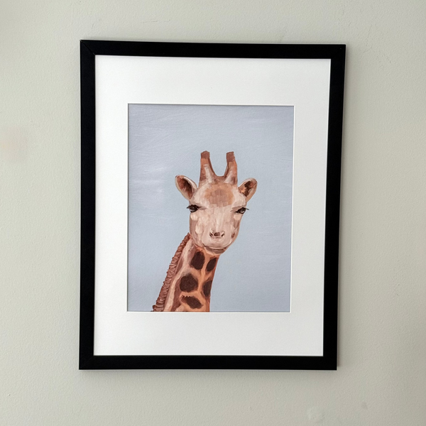 A framed art print featuring a painted giraffe head and neck against a pale blue-gray background, mounted on a light beige wall. The print has a white mat and is enclosed in a slim black frame.