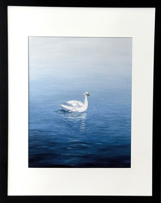 A framed art print featuring a white swan swimming on a calm blue lake, mounted on a light beige wall. The print has a white mat and is enclosed in a simple black frame.