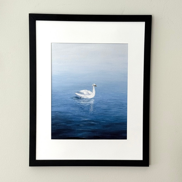 A framed art print featuring a white swan swimming on a calm blue lake, mounted on a light beige wall. The print has a white mat and is enclosed in a simple black frame.