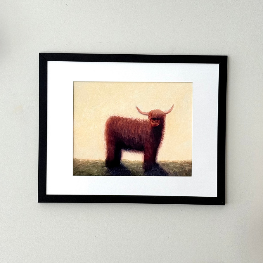 A framed art print featuring a brown Highland cow illustration on a cream background, set in a black frame with a white mat against a light gray wall.