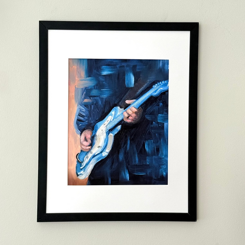 A framed art print featuring an expressive blue painting of a person playing an electric guitar, mounted on a light beige wall.