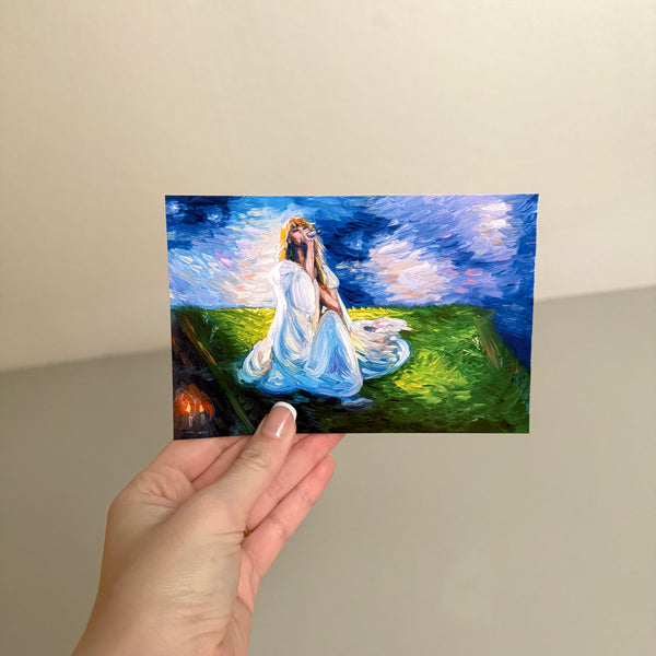 A hand holds a small rectangular glossy postcard depicting a woman in a flowing white dress seated on a grassy hill under a dramatic blue sky. The background is a plain beige wall.