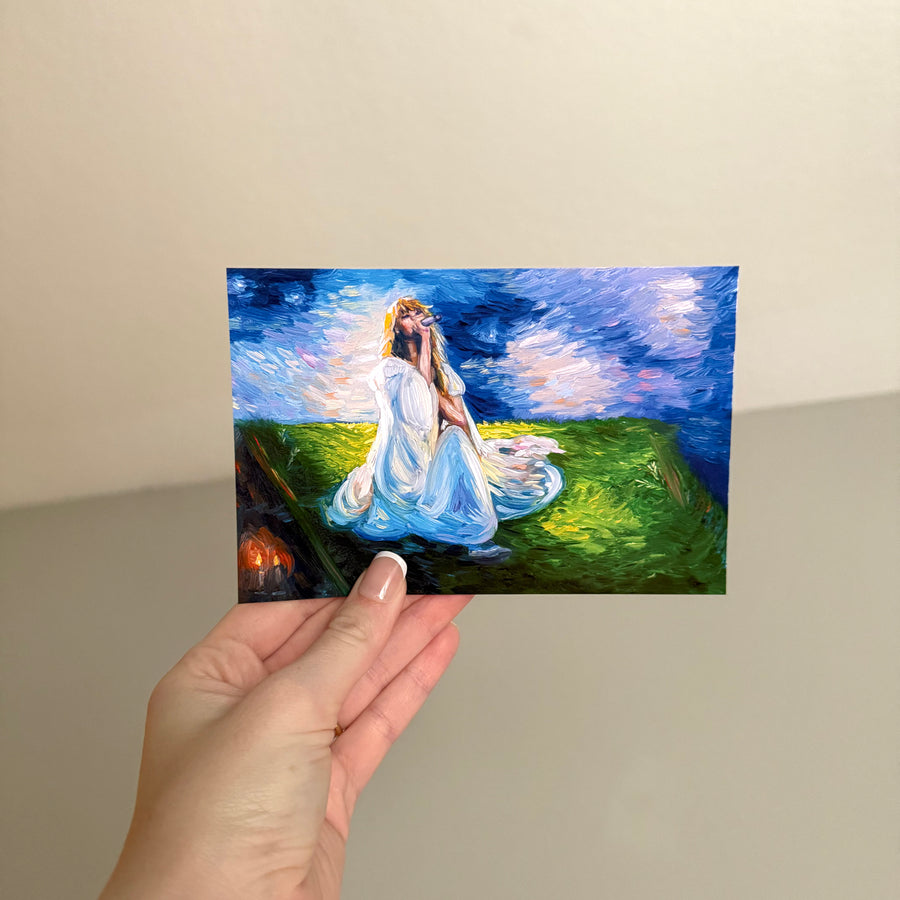 A hand holds a small rectangular glossy postcard depicting a woman in a flowing white dress seated on a grassy hill under a dramatic blue sky. The background is a plain beige wall.