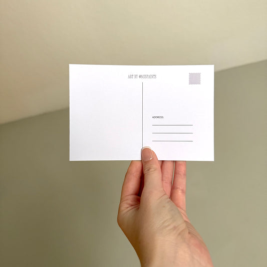 A hand holding a white postcard with minimalist black text and a small gray stamp box, set against a soft beige background.