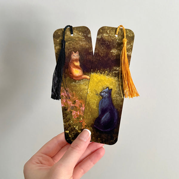 Set of two bookmarks featuring a grey and orange cat and tassels held by a hand against a beige background