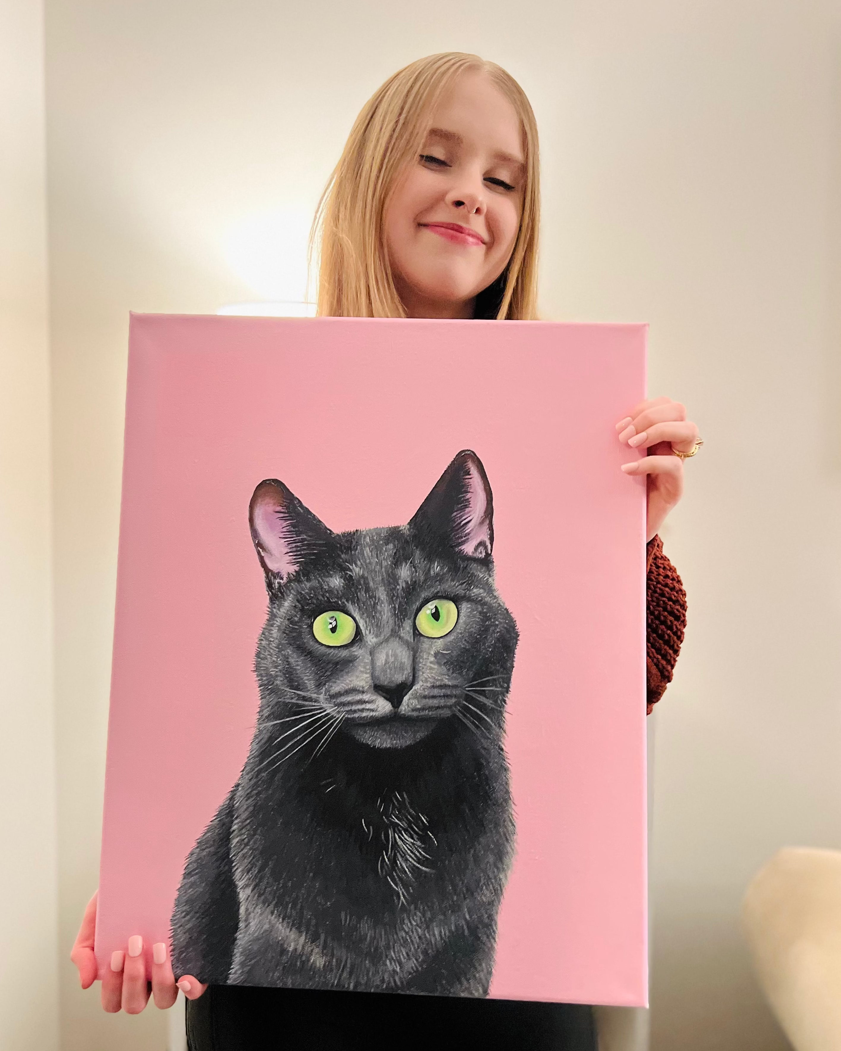 Person holding a painting of a gray cat with green eyes on a pink background