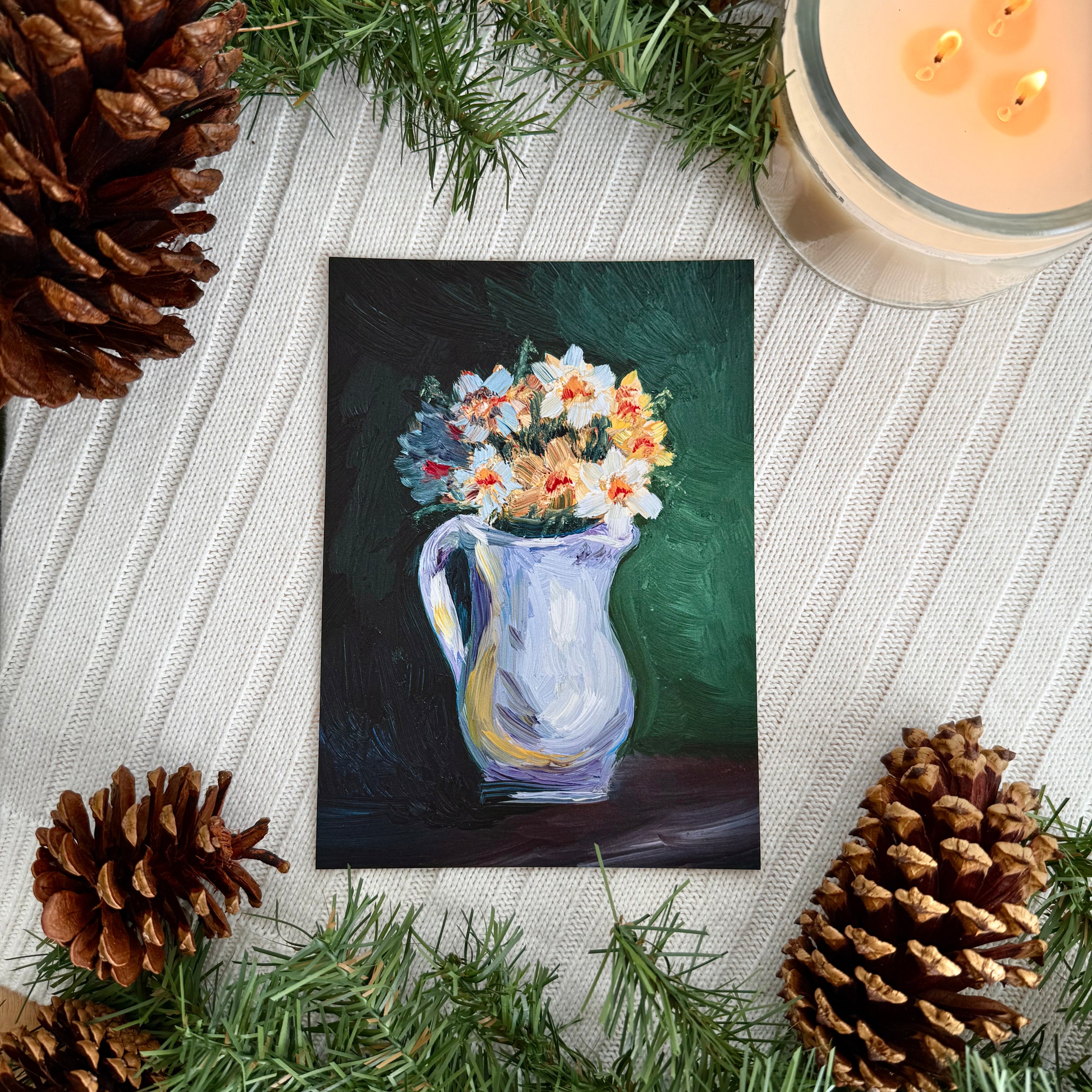 Print of a vase of daffodils on a knitted surface surrounded by evergreen branches, pinecones, and a candle