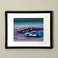 Print of a painting of two race cars in a black frame against a white wall.
