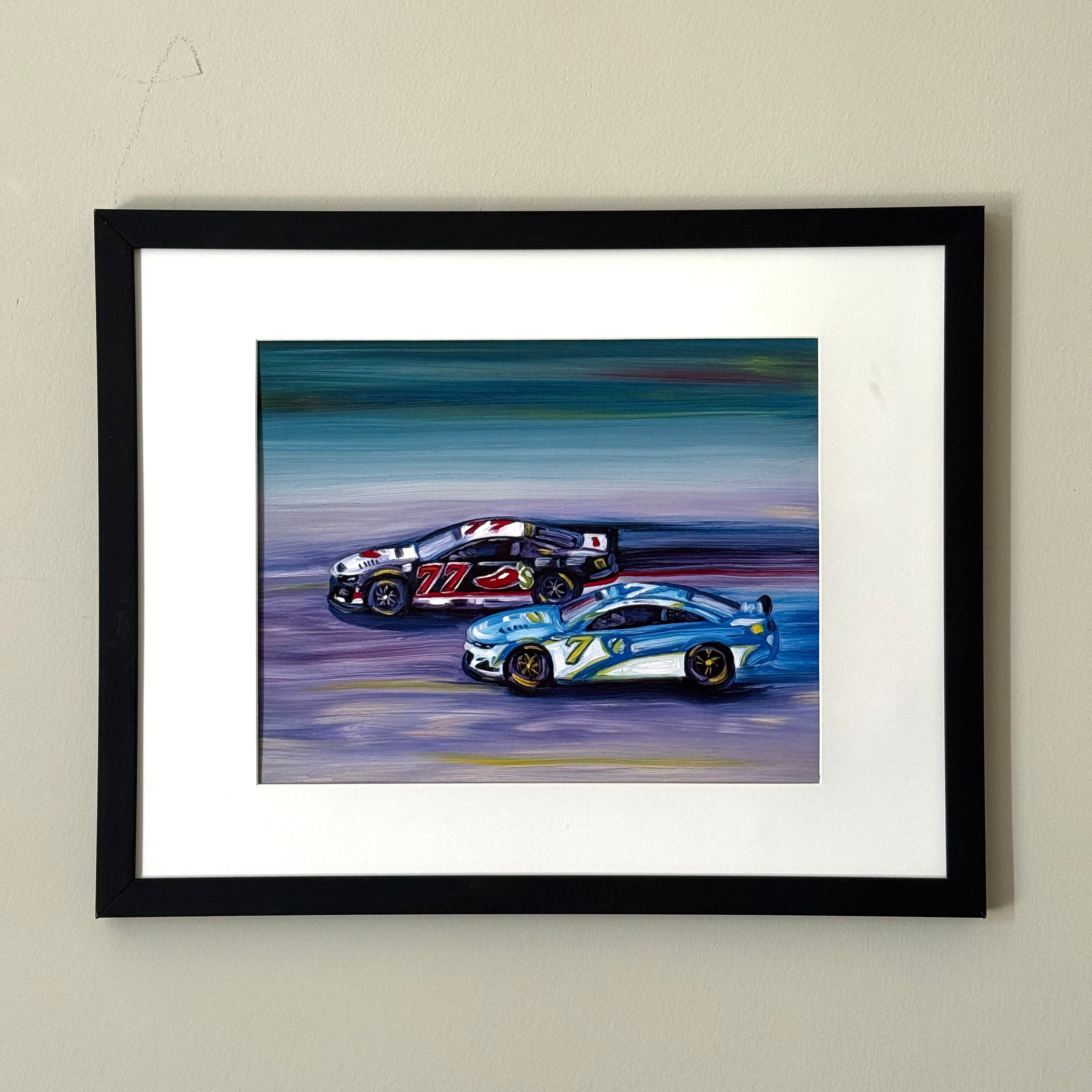 Print of a painting of two race cars in a black frame against a white wall.