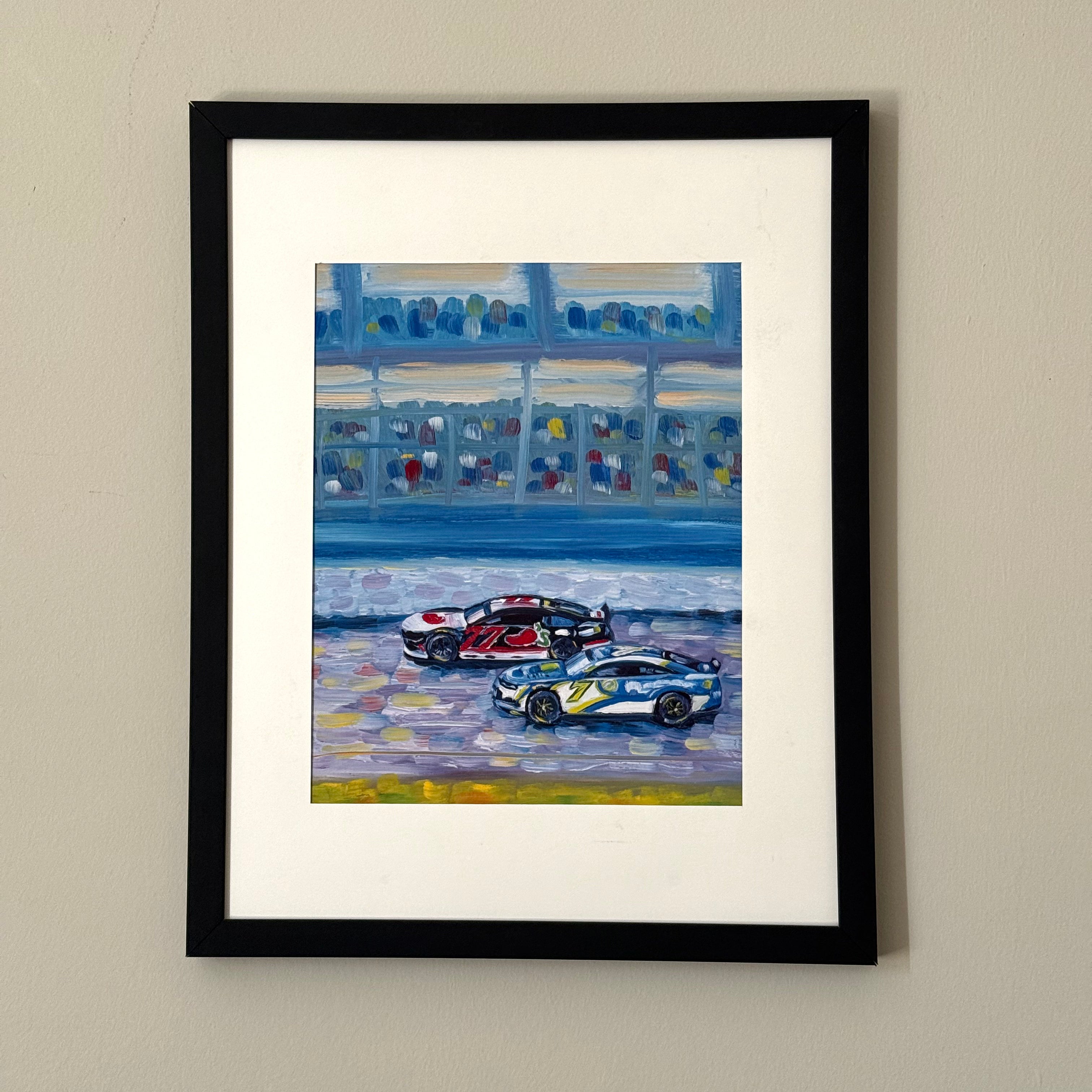 Print of a painting of two race cars in a black frame against a white wall.