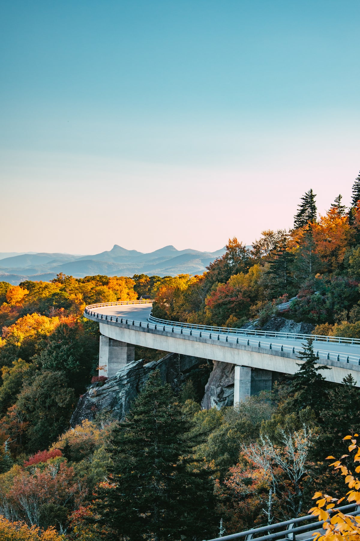 A scenic landscape photograph of a curved highway bridge spanning a rocky mountain pass, surrounded by autumn-colored forests and distant blue mountains under a clear sky.