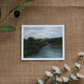 Burlap background with various floral decorations surrounding a fine art print depicting a river scene with dark blue water, green trees, and a white cloudy sky
