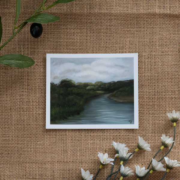 Burlap background with various floral decorations surrounding a fine art print depicting a river scene with dark blue water, green trees, and a white cloudy sky