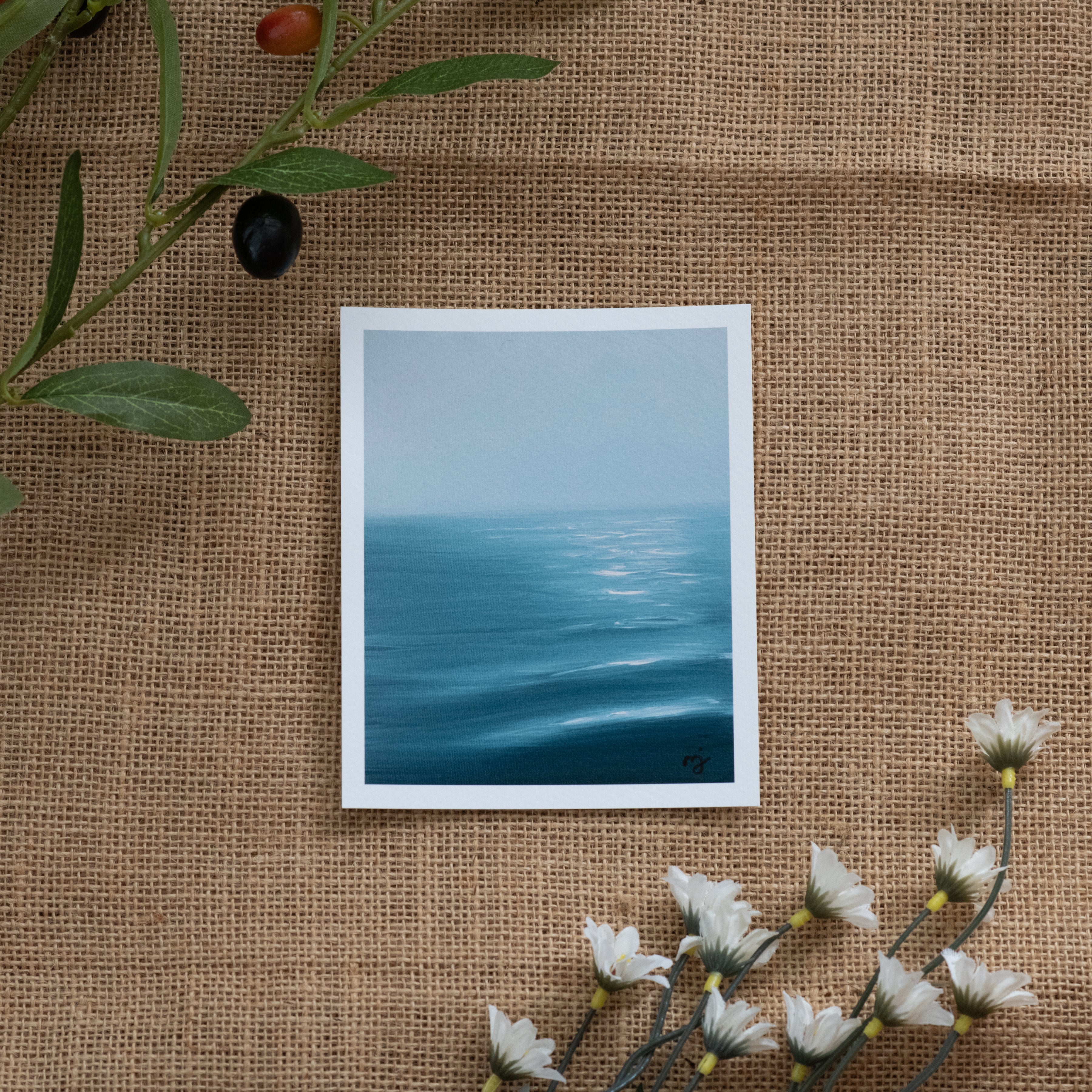 Burlap background with various floral decorations surrounding a fine art print depicting blue ocean waves with sun reflections and a light blue sky