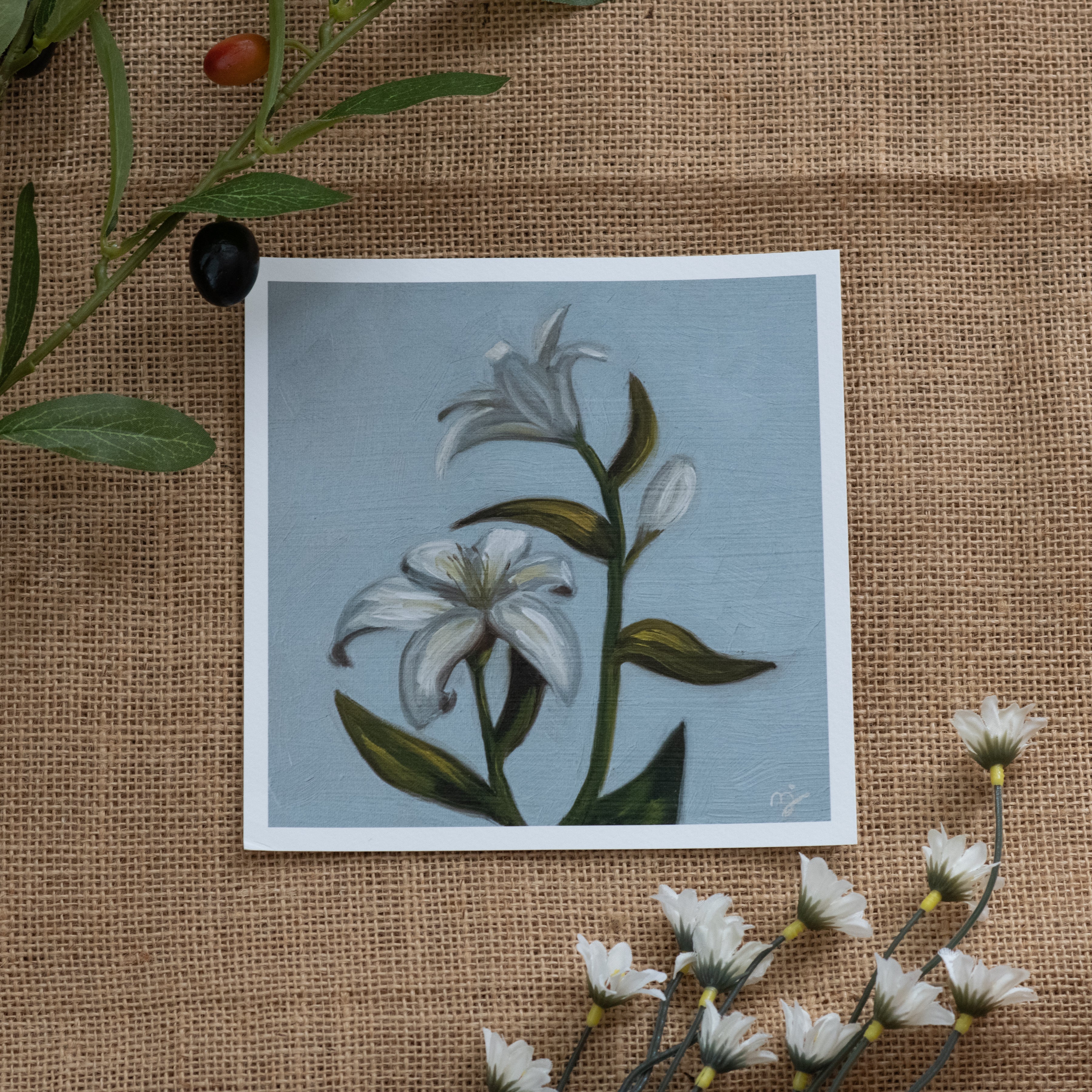 Burlap background with various floral decorations surrounding a fine art print depicting white lilies on a blue background