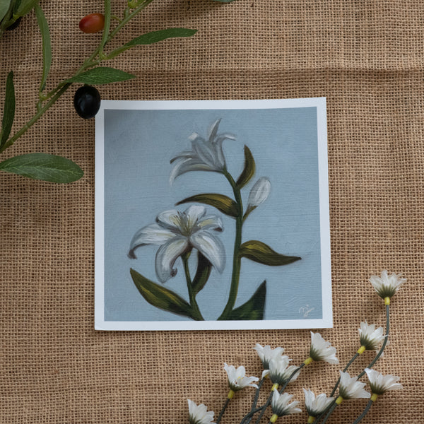 Burlap background with various floral decorations surrounding a fine art print depicting white lilies on a blue background