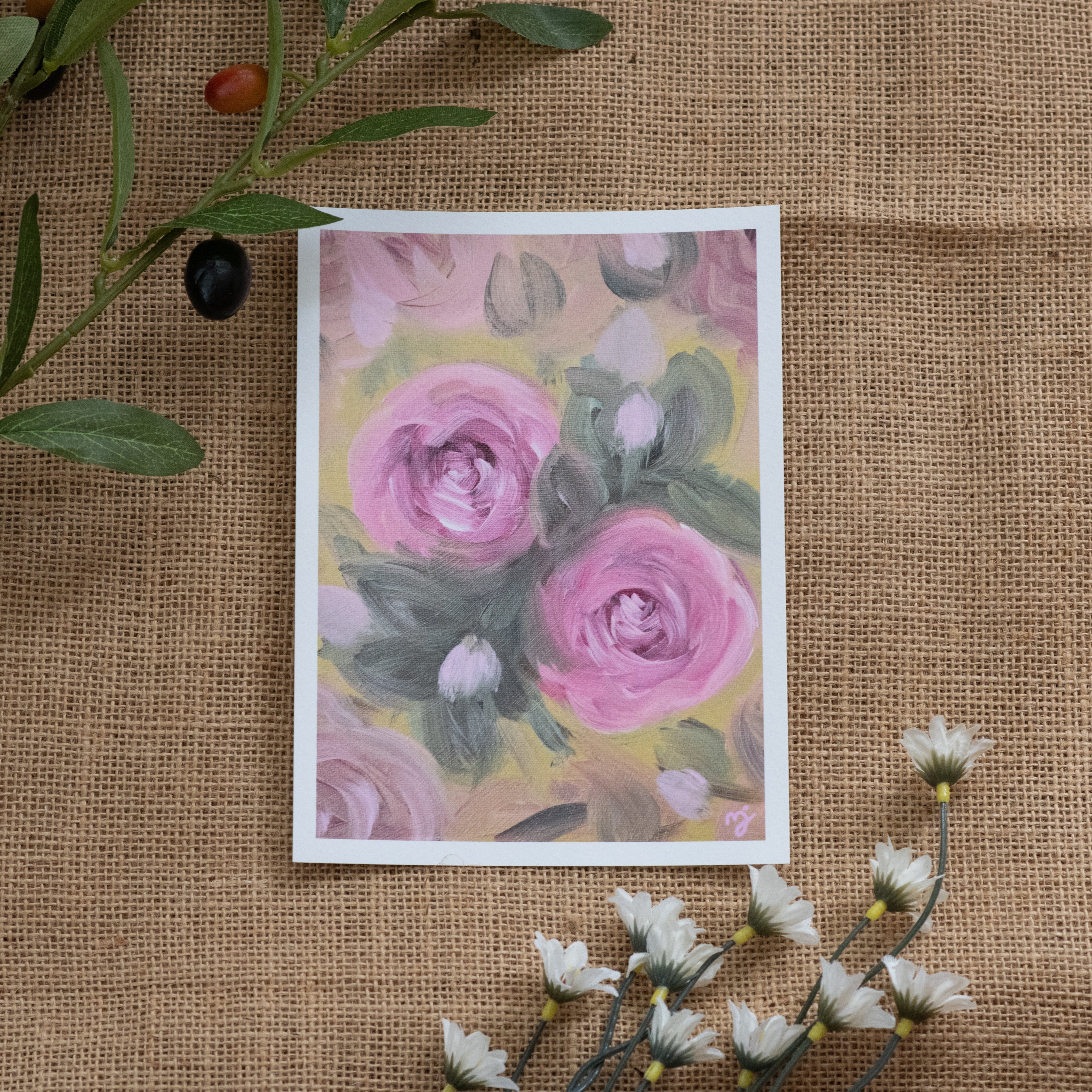 Burlap background with various floral decorations surrounding a fine art print depicting pink roses on a yellow background
