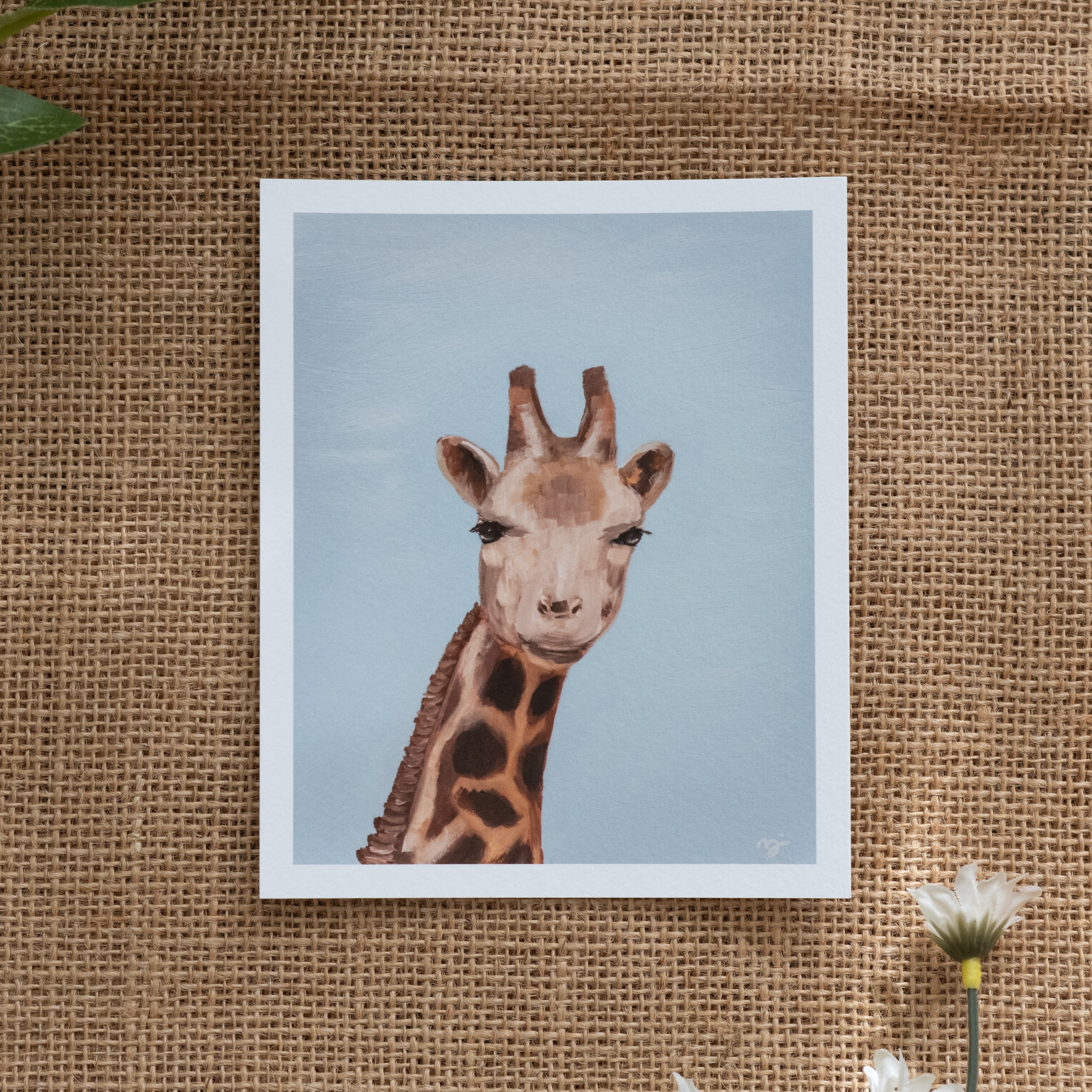 Burlap background with various floral decorations surrounding a fine art print depicting a giraffe's head and neck against a blue sky