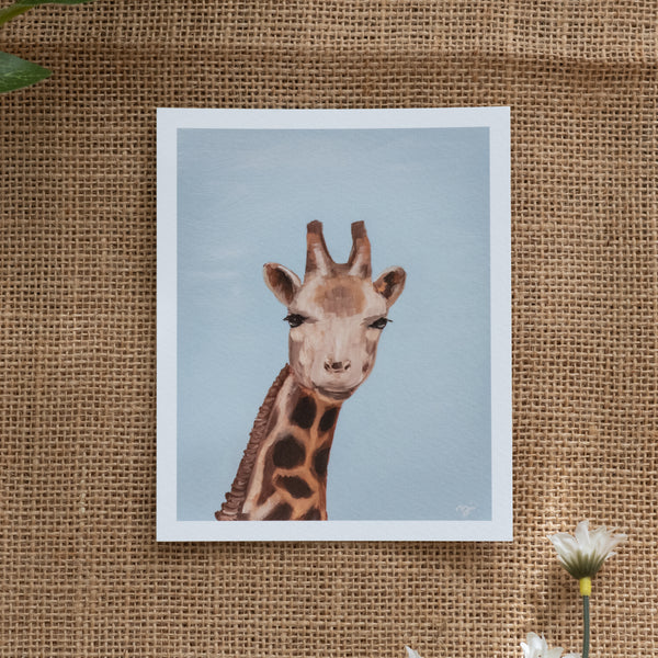 Burlap background with various floral decorations surrounding a fine art print depicting a giraffe's head and neck against a blue sky