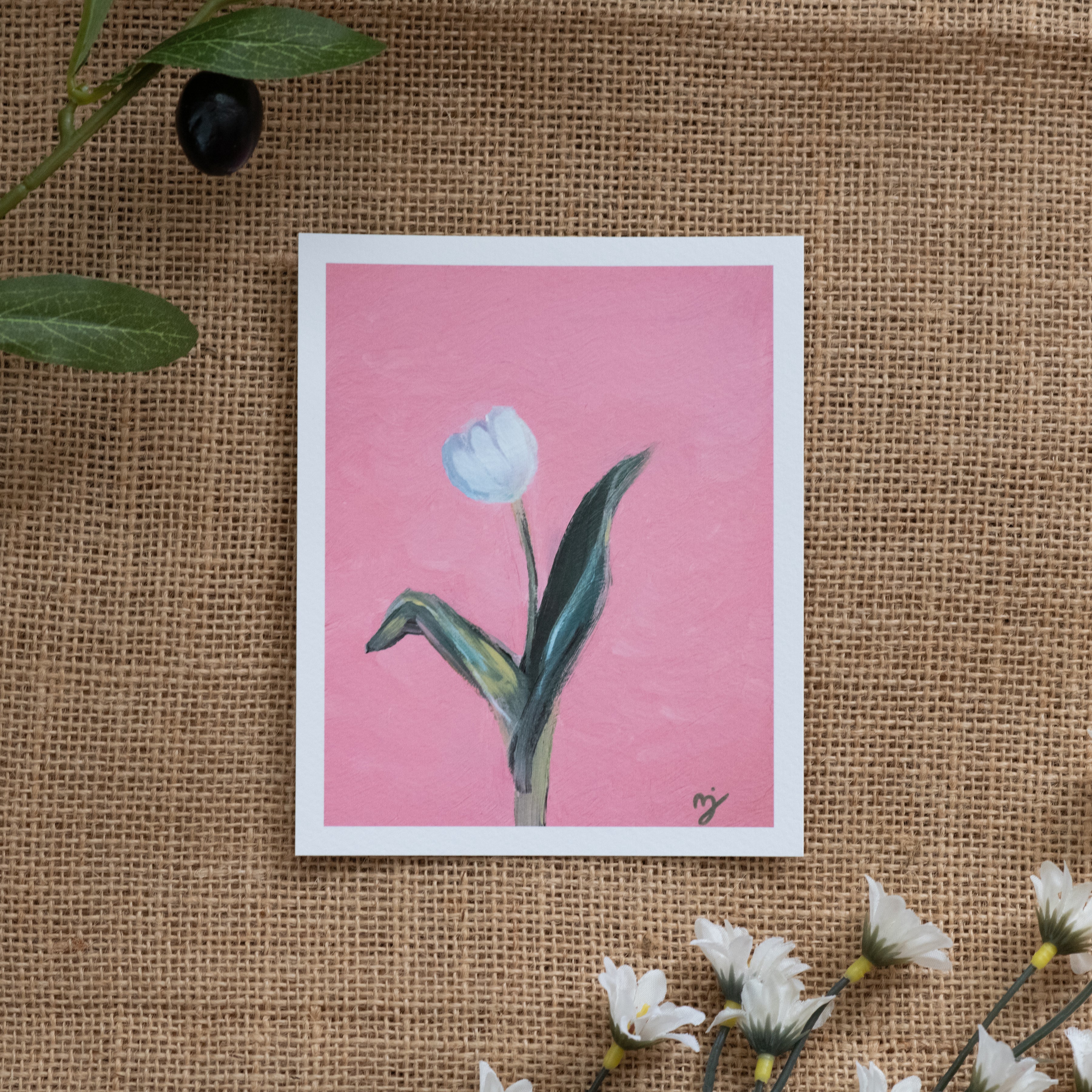 Burlap background with various floral decorations surrounding a fine art print depicting a white tulip on a pink background