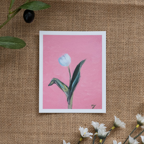 Burlap background with various floral decorations surrounding a fine art print depicting a white tulip on a pink background