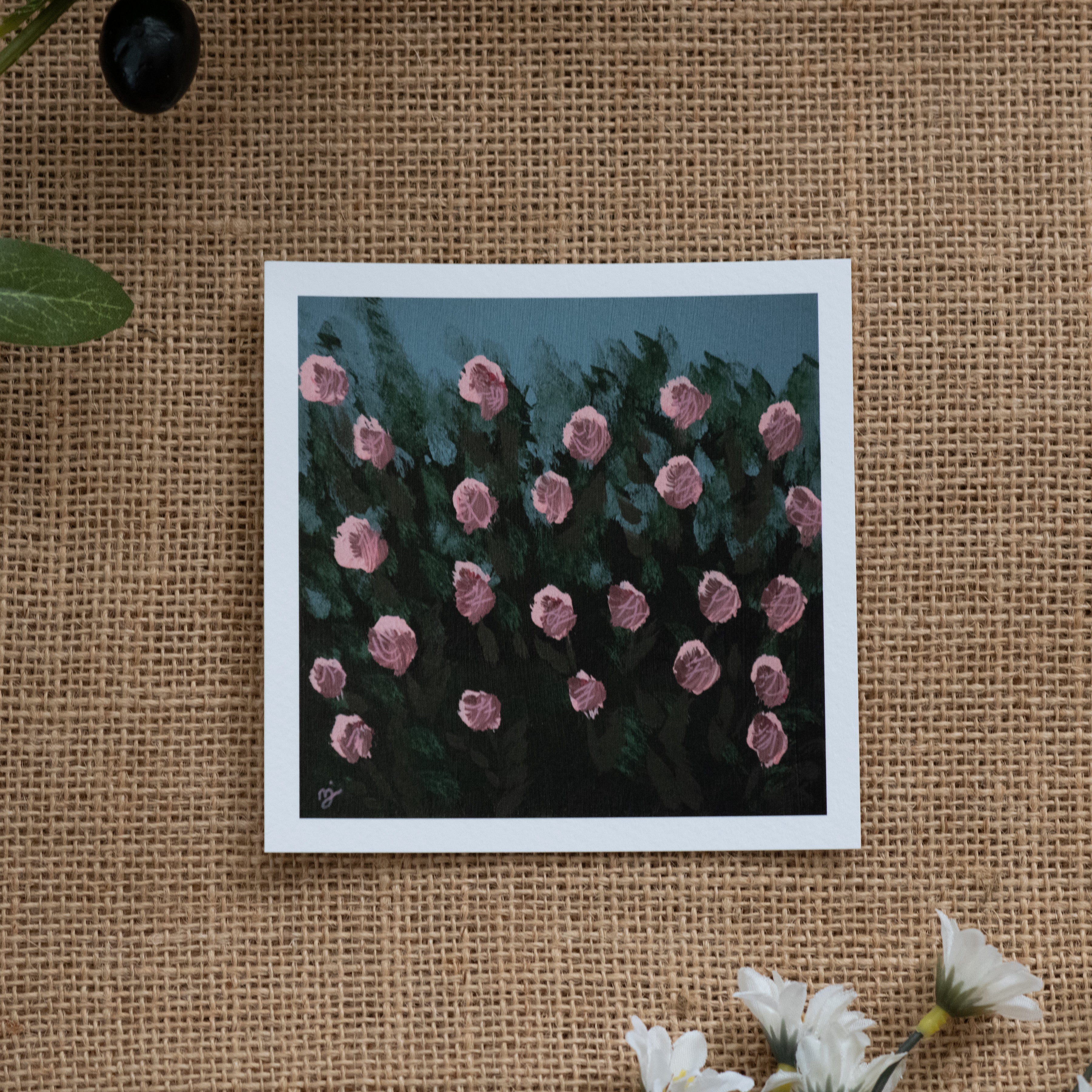 Burlap background with various floral decorations surrounding a fine art print depicting a bunch of pink roses and green shrubbery with a blue sky