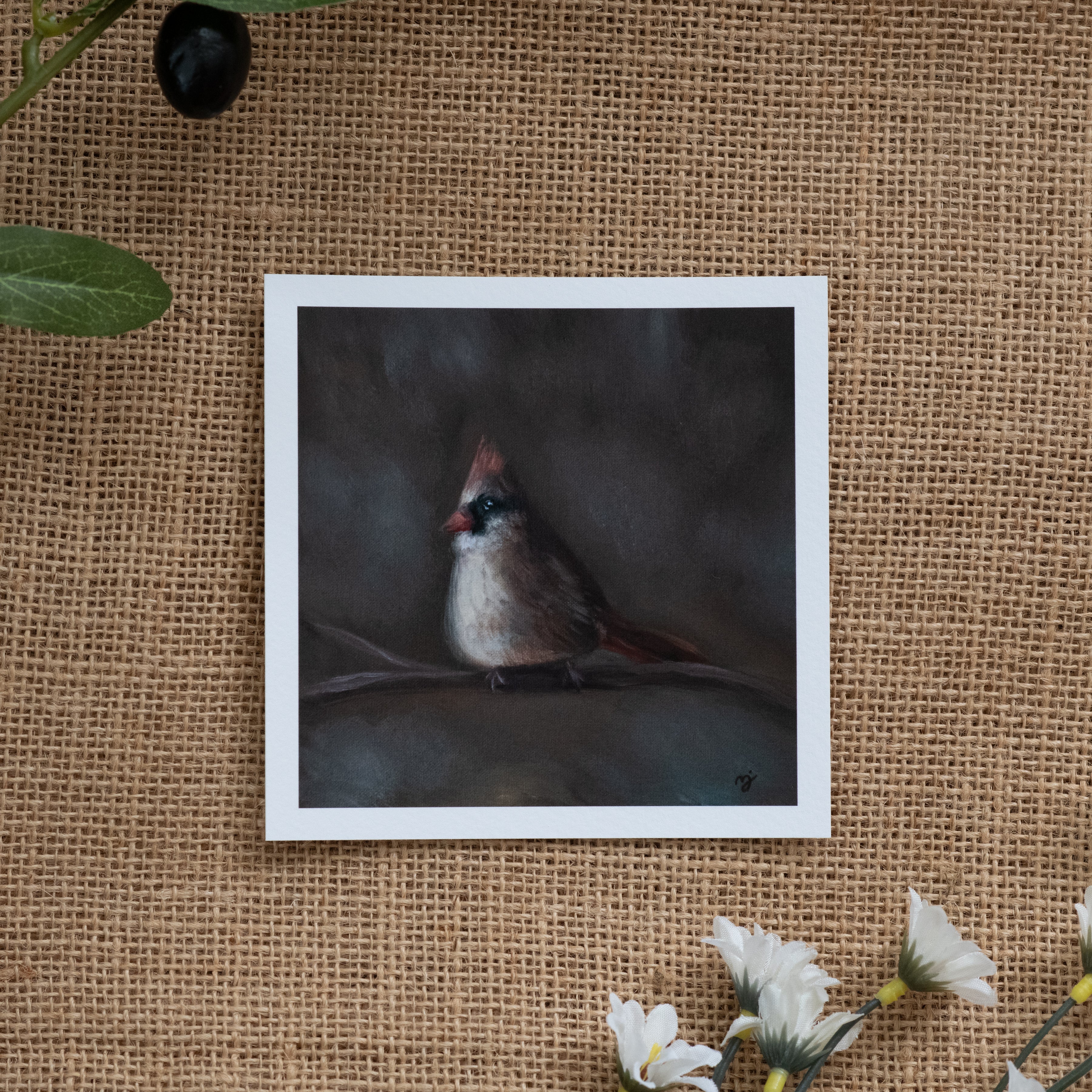 Burlap background with various floral decorations surrounding a fine art print depicting a female cardinal sitting on a branch with blurred brown grey background 