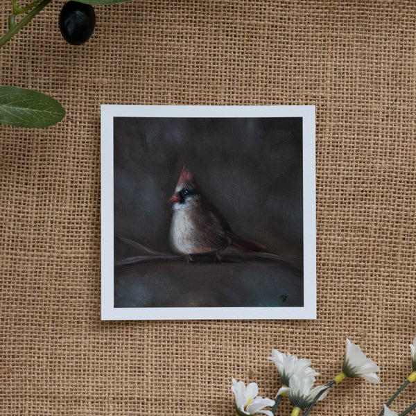 Burlap background with various floral decorations surrounding a fine art print depicting a female cardinal sitting on a branch with blurred brown grey background 