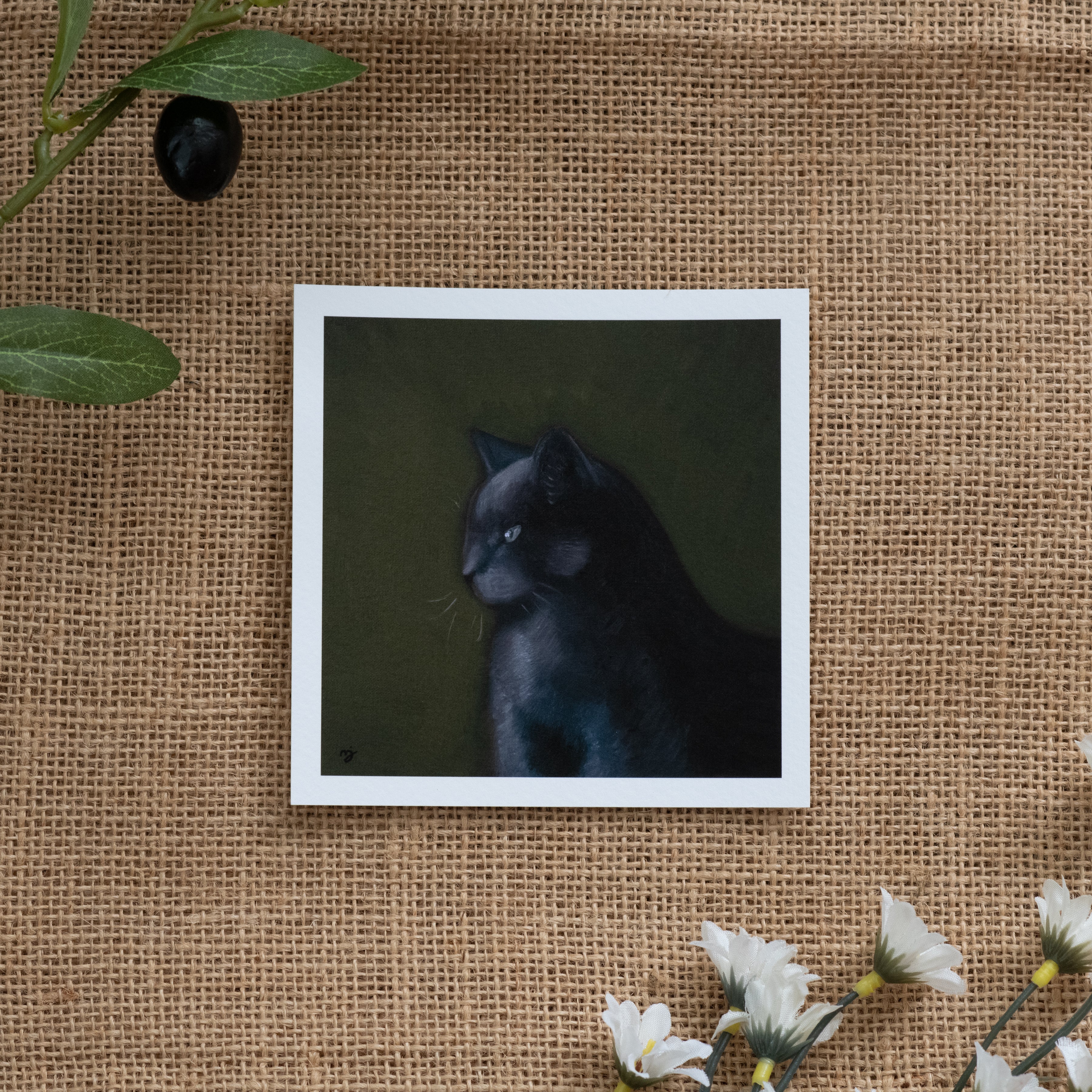Burlap background with various floral decorations surrounding a fine art print depicting a dark grey cat against a green background